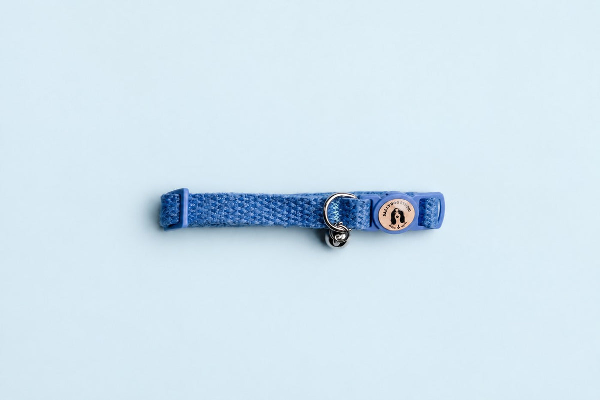 Salt Dog Studios cornflower blue hemp cat collar with matching breakaway buckle, logo and silver bell on a pale blue background.