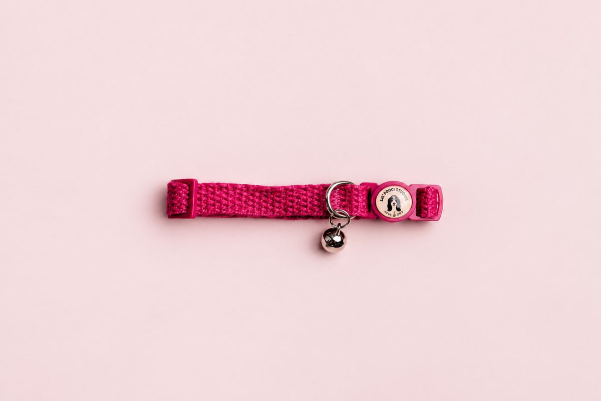 Salt Dog Studios magenta pink hemp cat collar with matching breakaway buckle, logo and silver bell on a rose blush background.