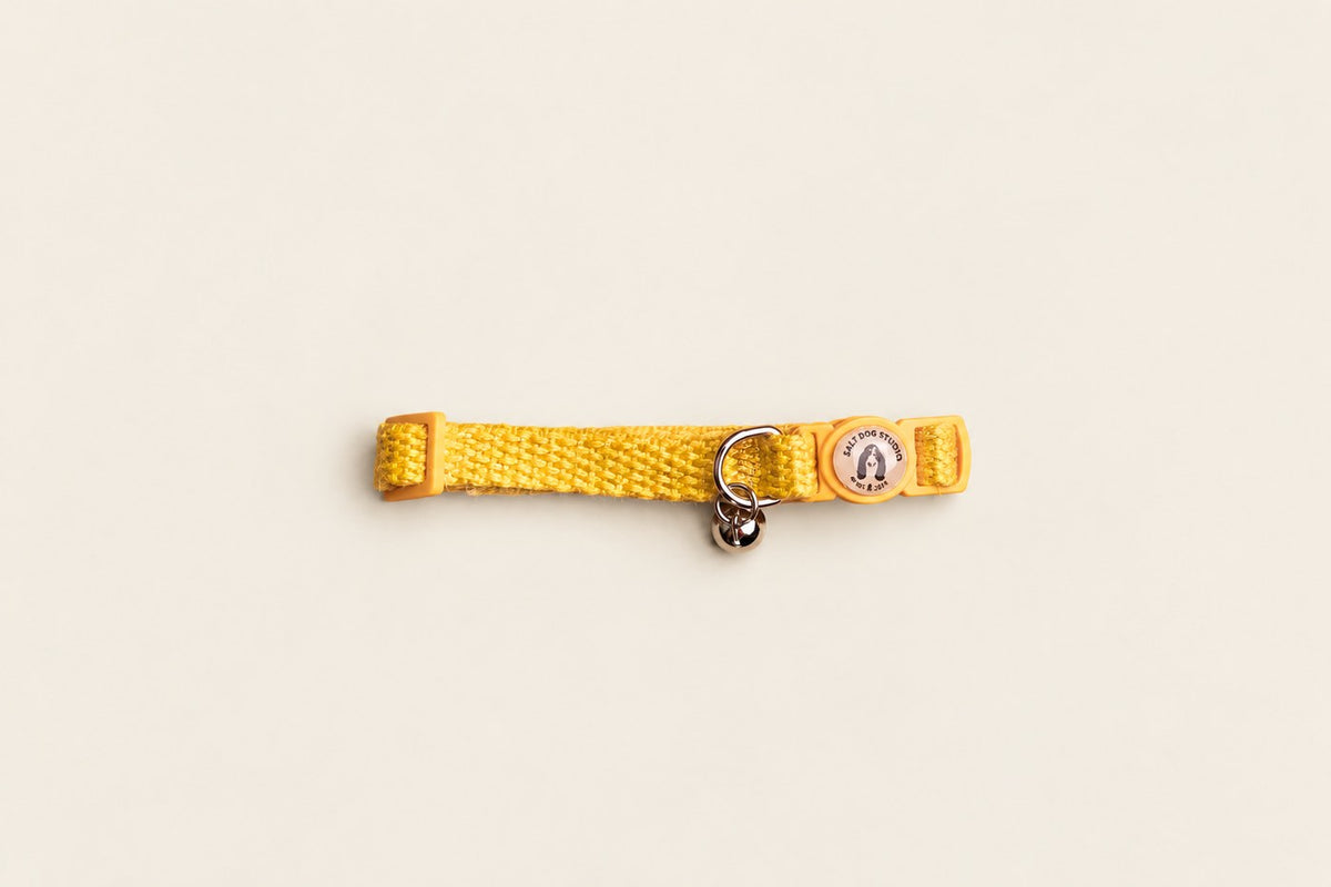 Salt Dog Studios sunflower yellow hemp cat collar with matching breakaway buckle, logo and silver bell on a pale beige background.